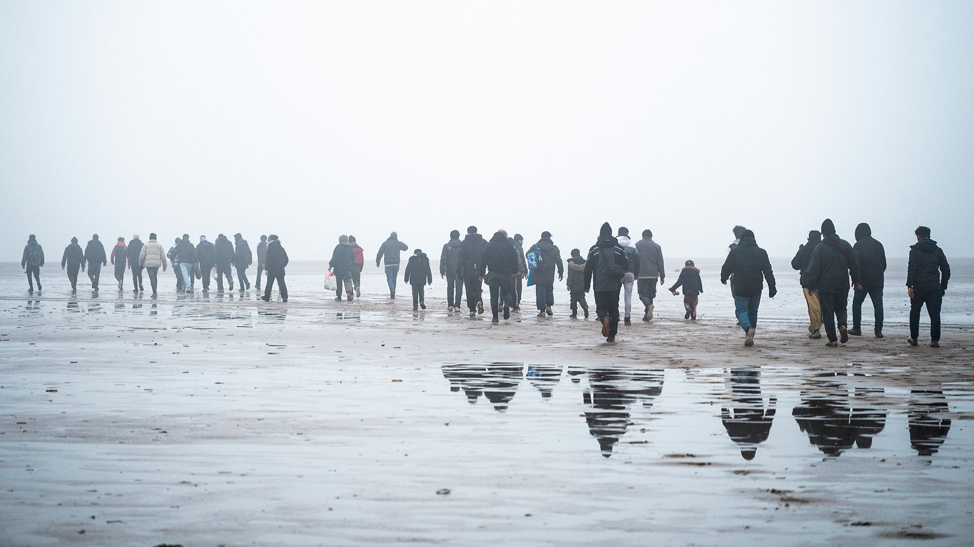 Un grupo de personas migrantes camina por la playa de Petit-Fort-Philippe, en Gravelines, Francia, el 17 de enero de 2025, mientras se preparan para partir en barco hacia el Reino Unido. Las personas, de espaldas a la cámara, avanzan hacia la embarcación en un día gris, con niebla y cielo nublado. Europa