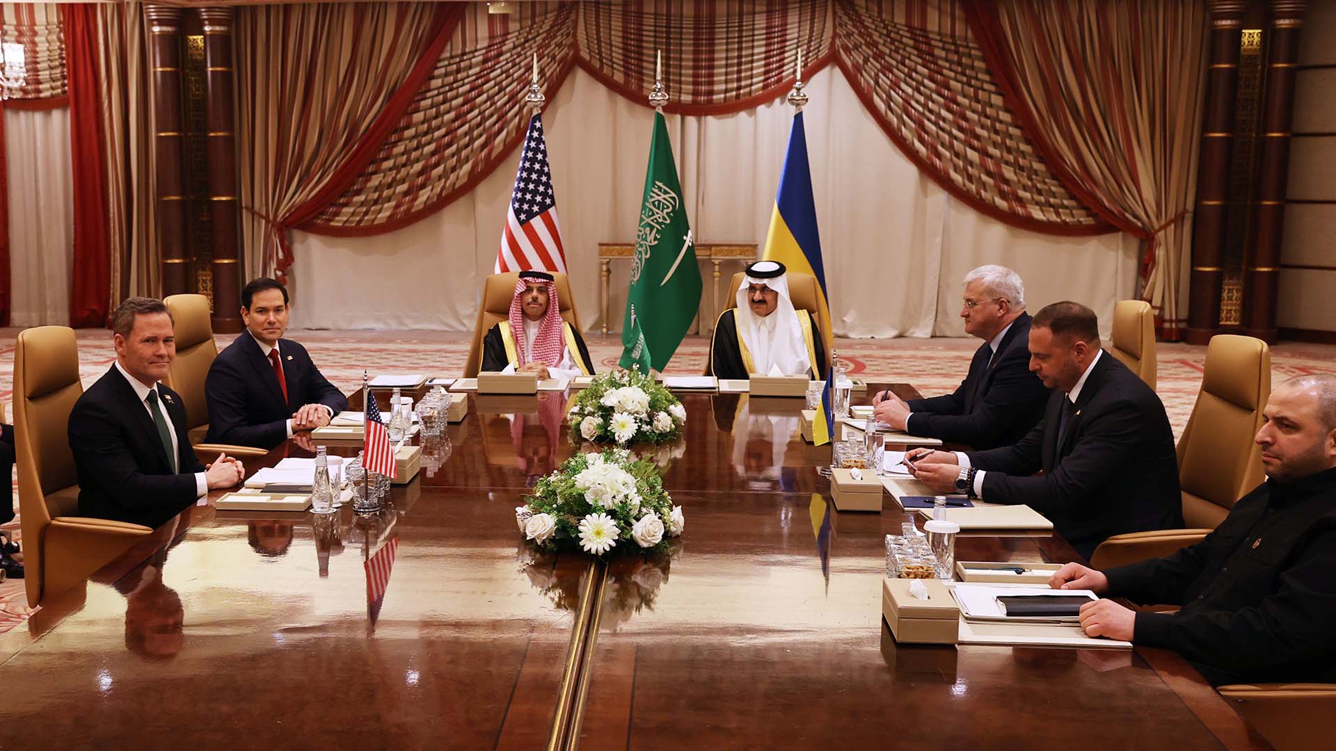 De izquierda a derecha, sentados alrededor de una mesa de madera: el asesor de Seguridad Nacional de EEUU, Mike Waltz; el secretario de Estado de EEUU, Marco Rubio; el ministro de Asuntos Exteriores de Arabia Saudí, Faisal bin Farhan; el asesor de Seguridad Nacional saudí, Mosaad bin Mohammad al-Aiban; el ministro de Asuntos Exteriores de Ucrania, Andrii Sybiha; el jefe de la Oficina Presidencial de Ucrania, Andriy Yermak, y el ministro de Defensa de Ucrania, Rustem Umerov, asisten a una reunión entre EEUU y Ucrania organizada por Arabia Saudí el 11 de marzo de 2025 en Yeda. La delegación estadounidense se encuentra a la izquierda, los representantes saudíes en la cabecera y los ucranianos a la derecha. En el fondo se aprecian las banderas de los tres países representadas. Ucrania