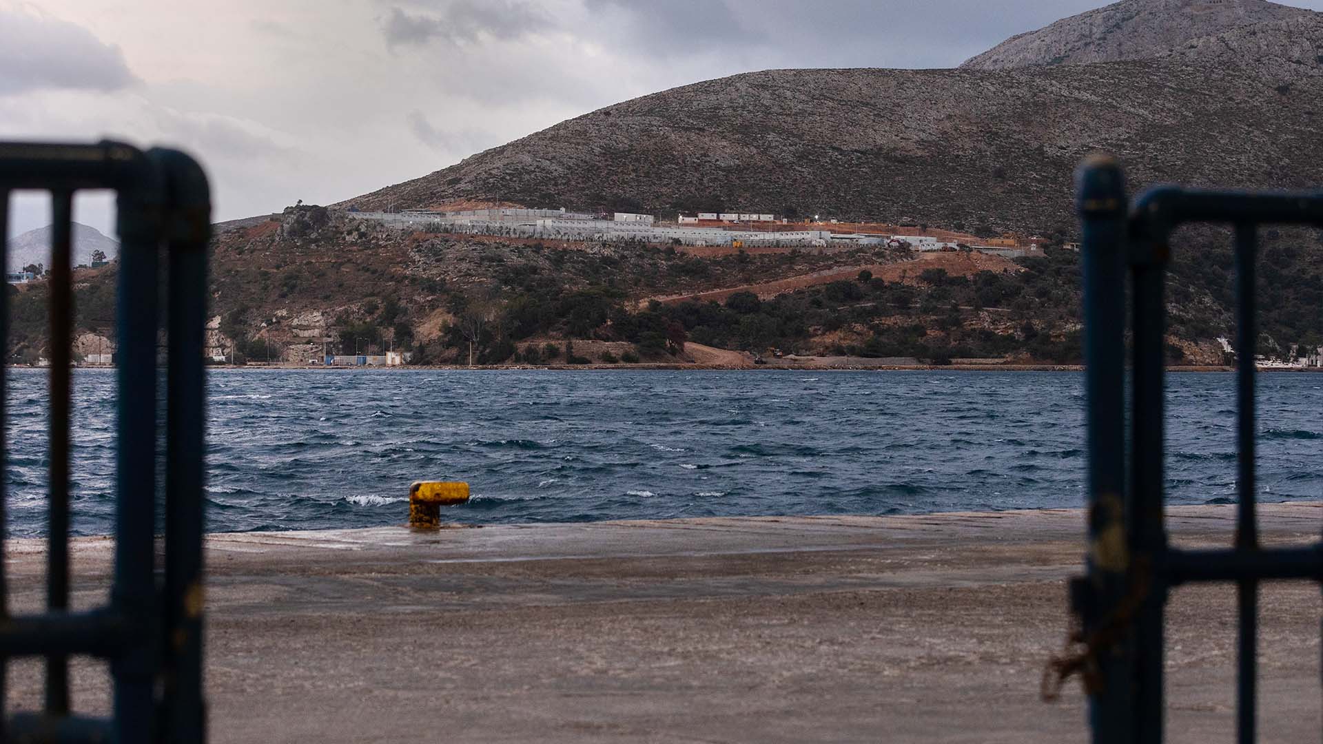 Vista del Centro de Recepción e Identificación de usos múltiples cerca de Lakki, en la isla de Leros (Grecia). El centro, rodeado por una valla de seguridad, se encuentra en una colina, desde donde se divisa el mar en primer plano y un cielo nublado al fondo. Migración