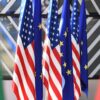 Varias banderas de Estados Unidos y la Unión Europea colocadas en soportes dorados sobre una mesa azul durante la cumbre UE-EEUU de 2017. En primer plano, parcialmente desenfocadas, se ven otras banderas con colores rojo y verde. Europa