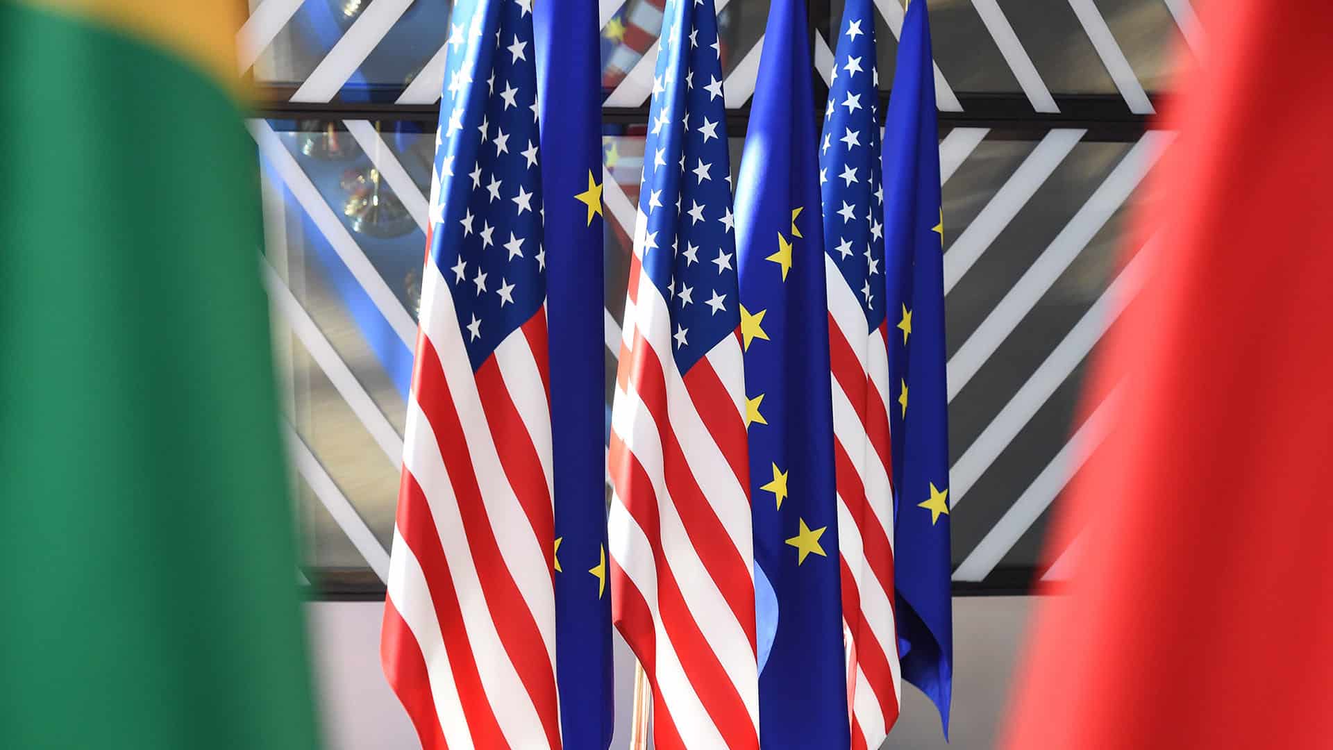 Varias banderas de Estados Unidos y la Unión Europea colocadas en soportes dorados sobre una mesa azul durante la cumbre UE-EEUU de 2017. En primer plano, parcialmente desenfocadas, se ven otras banderas con colores rojo y verde. Europa