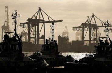 Siluetas de grúas pórtico y barcos de remolque en la terminal 'Eurogate' del puerto de Hamburgo al atardecer, destacándose contra un cielo nublado en tonos ocre. En primer plano, varios remolcadores reposan en el agua, mientras al fondo se perciben contenedores y estructuras portuarias. Trump