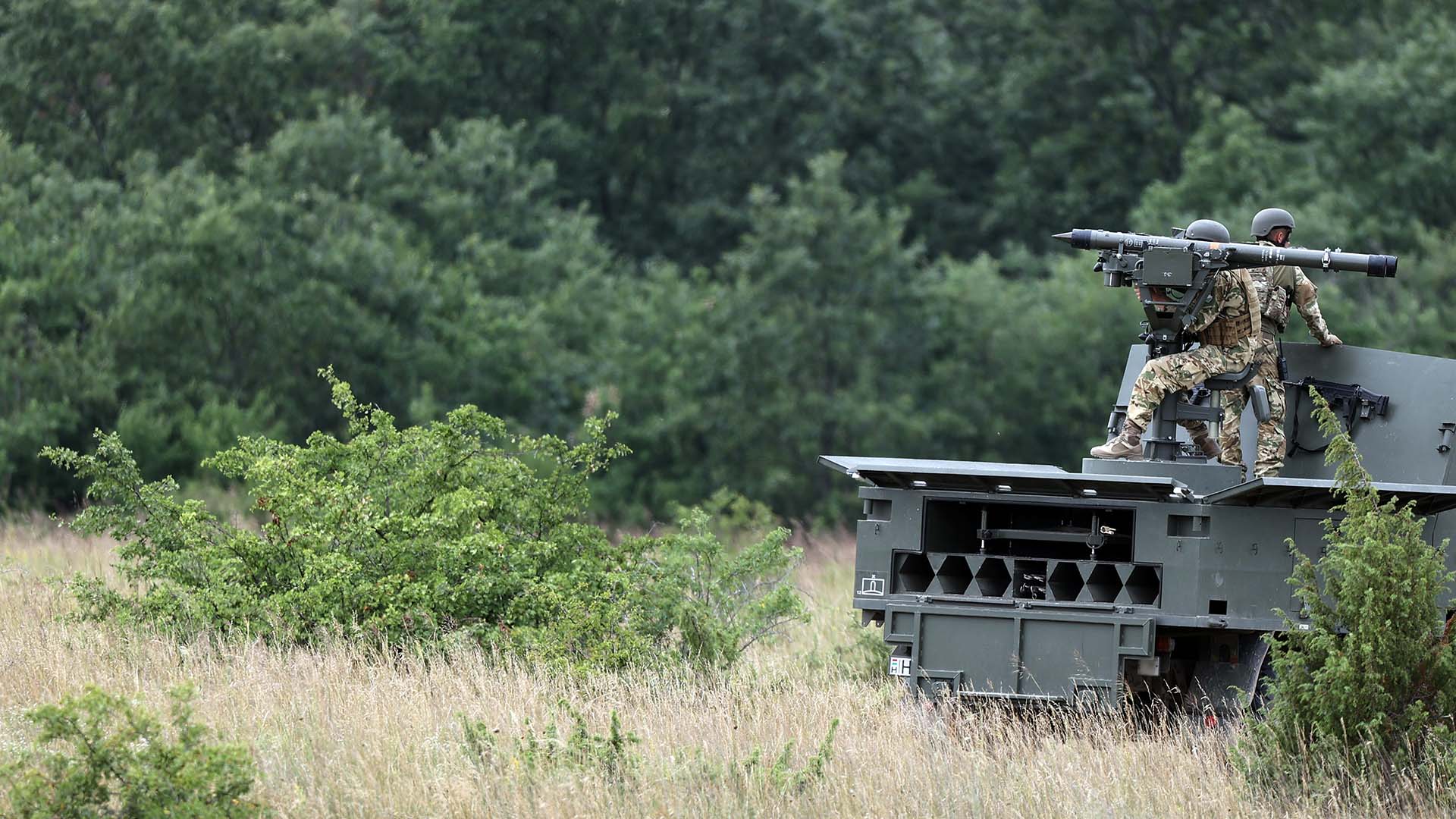 Dos soldados con uniforme de combate operan un sistema de misiles antiaéreos sobre un vehículo militar durante el ejercicio de helicópteros Fire Blade 2022 de la Agencia Europea de Defensa en Hungría. En el fondo, se aprecia el follaje verde del campo abierto donde se desarrolla la maniobra. Libro Blanco