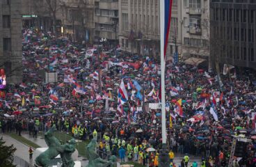 Multitud de manifestantes marchando en Belgrado (Serbia), con banderas y pancartas en protesta contra la corrupción gubernamental, mientras la policía y personal de seguridad supervisan el evento. Serbia