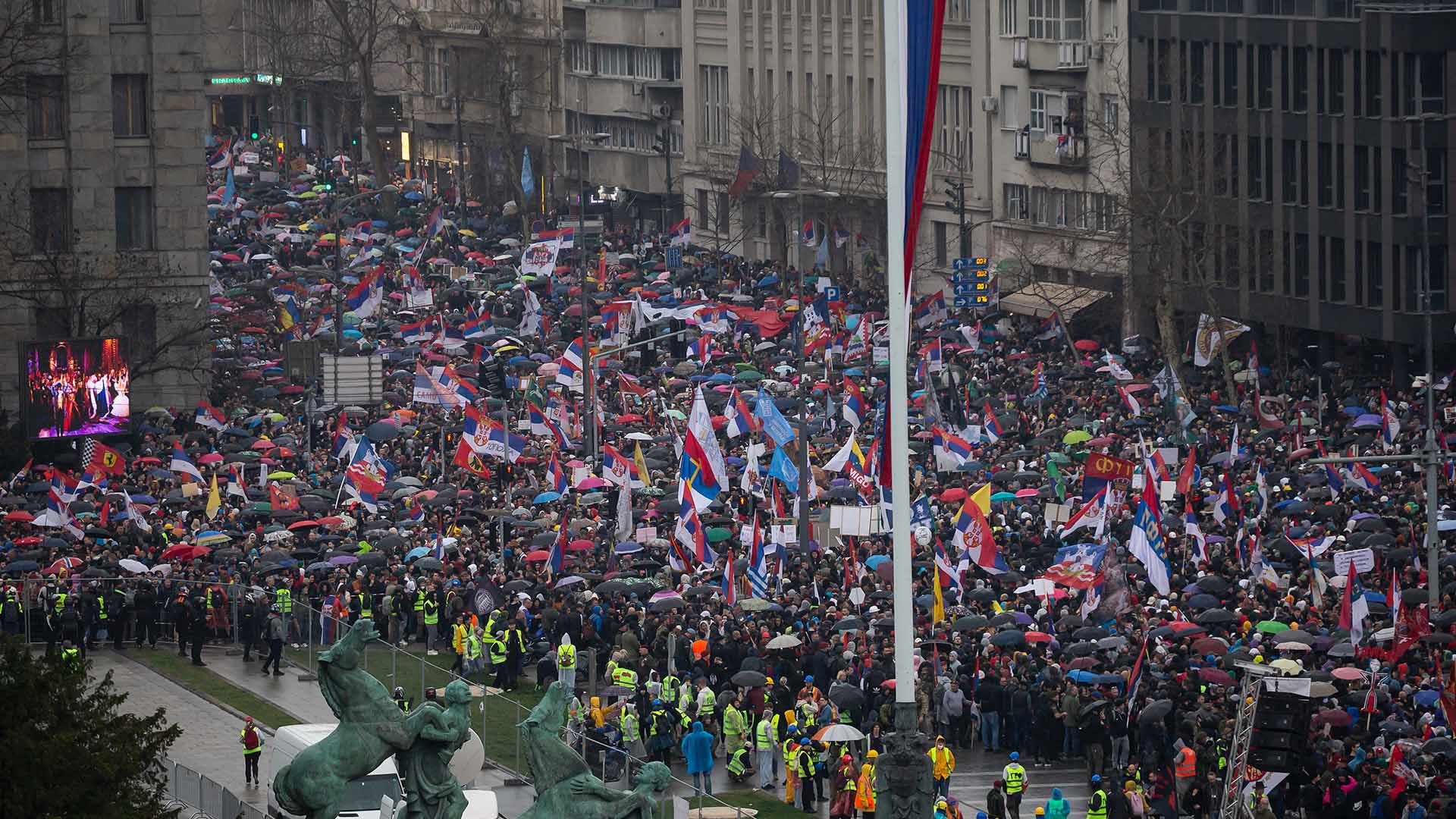 Multitud de manifestantes marchando en Belgrado (Serbia), con banderas y pancartas en protesta contra la corrupción gubernamental, mientras la policía y personal de seguridad supervisan el evento. Serbia