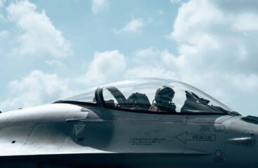 A Belgian F-16 pilot, wearing a flight helmet, oxygen mask, and full flight suit, sits inside the bubble canopy of a grey F-16 Fighting Falcon. The cockpit features visible instrument panels and side-stick controls, with warning labels on the fuselage just below the cockpit. The aircraft is parked on the tarmac at Florennes Air Base under a bright, partly cloudy sky during a NATO Quick Reaction Alert (QRA) exercise. Defence