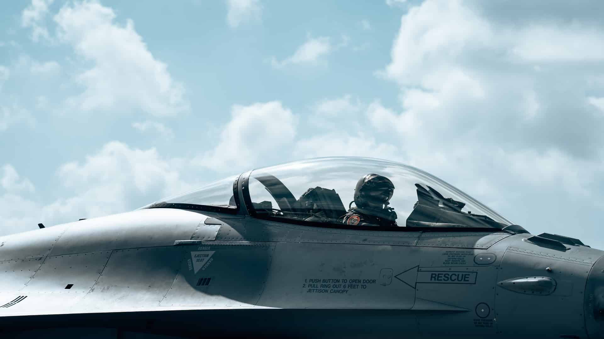 A Belgian F-16 pilot, wearing a flight helmet, oxygen mask, and full flight suit, sits inside the bubble canopy of a grey F-16 Fighting Falcon. The cockpit features visible instrument panels and side-stick controls, with warning labels on the fuselage just below the cockpit. The aircraft is parked on the tarmac at Florennes Air Base under a bright, partly cloudy sky during a NATO Quick Reaction Alert (QRA) exercise. Defence