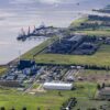 Vista diurna de la terminal flotante de GNL en Brunsbüttel (Alemania), donde el mar se encuentra con extensas áreas verdes. Se observa un puerto con buques y grúas, una planta industrial, turbinas eólicas y una subestación eléctrica, parte de la infraestructura energética del país. Rusia