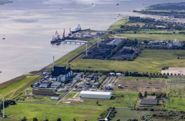 Vista diurna de la terminal flotante de GNL en Brunsbüttel (Alemania), donde el mar se encuentra con extensas áreas verdes. Se observa un puerto con buques y grúas, una planta industrial, turbinas eólicas y una subestación eléctrica, parte de la infraestructura energética del país. Rusia