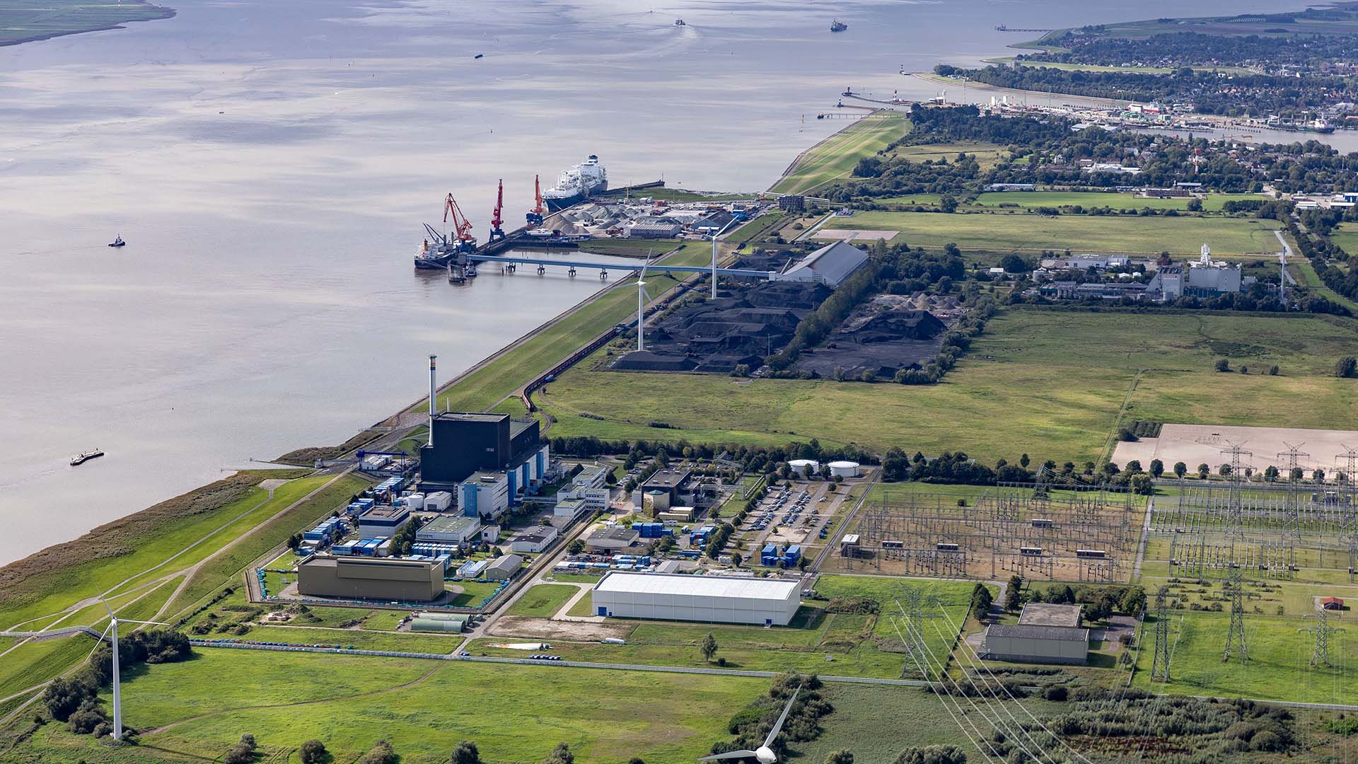 Vista diurna de la terminal flotante de GNL en Brunsbüttel (Alemania), donde el mar se encuentra con extensas áreas verdes. Se observa un puerto con buques y grúas, una planta industrial, turbinas eólicas y una subestación eléctrica, parte de la infraestructura energética del país. Rusia