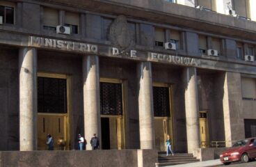 Fachada de la sede del Ministerio de Economía en Buenos Aires, Argentina. Tres personas hacen fila, mientras un policía les observa. Un hombre (con traje y maletín) y una mujer (que camina en sentido contrario) pasan por la acera.