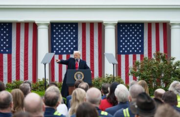 Donald Trump, desde un atril con micrófono, señala con su brazo derecho durante su alocución ante una audiencia congregada en el evento Make America Wealthy Again o “Día de la Liberación” en la Rosaleda de la Casa Blanca, el pasado 2 de abril de 2025. Detrás, cuatro banderas de EEUU cuelgan verticalmente entre los pilares de columnas.