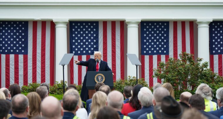 Donald Trump, desde un atril con micrófono, señala con su brazo derecho durante su alocución ante una audiencia congregada en el evento Make America Wealthy Again o “Día de la Liberación” en la Rosaleda de la Casa Blanca, el pasado 2 de abril de 2025. Detrás, cuatro banderas de EEUU cuelgan verticalmente entre los pilares de columnas.