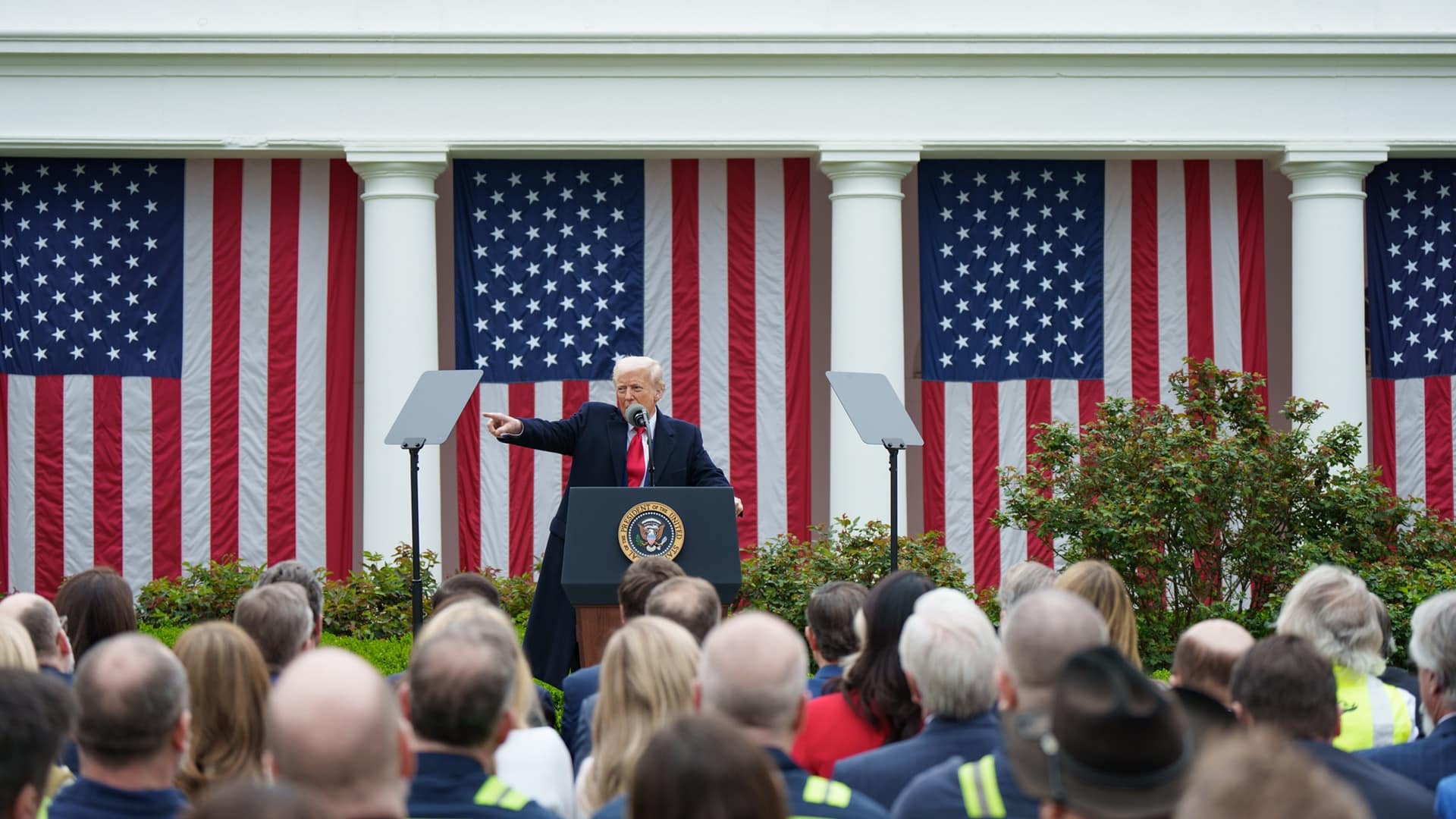 Donald Trump, desde un atril con micrófono, señala con su brazo derecho durante su alocución ante una audiencia congregada en el evento Make America Wealthy Again o “Día de la Liberación” en la Rosaleda de la Casa Blanca, el pasado 2 de abril de 2025. Detrás, cuatro banderas de EEUU cuelgan verticalmente entre los pilares de columnas.