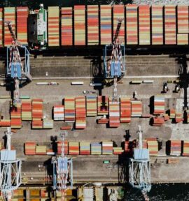 Vista aérea de un puerto marítimo industrial en Nápoles, con varias grúas de carga alineadas y contenedores de transporte multicolores organizados sobre el muelle y a bordo de un buque carguero atracado. UE