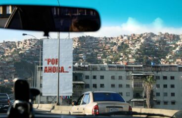 Foto de una pancarta blanca con la frase en rojo “Por ahora…”, tomada desde un coche en la autopista Caracas – La Guaira. Detrás de la pancarta, un edificio bajo de apartamentos y al fondo la imagen de un cerro o barriada popular en el oeste de la ciudad de Caracas (Venezuela). La frase fue pronunciada por Hugo Chávez Frías en su rendición ante los medios de comunicación, tras el intento de golpe de Estado en 1992.