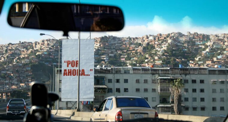 Foto de una pancarta blanca con la frase en rojo “Por ahora…”, tomada desde un coche en la autopista Caracas – La Guaira. Detrás de la pancarta, un edificio bajo de apartamentos y al fondo la imagen de un cerro o barriada popular en el oeste de la ciudad de Caracas (Venezuela). La frase fue pronunciada por Hugo Chávez Frías en su rendición ante los medios de comunicación, tras el intento de golpe de Estado en 1992.