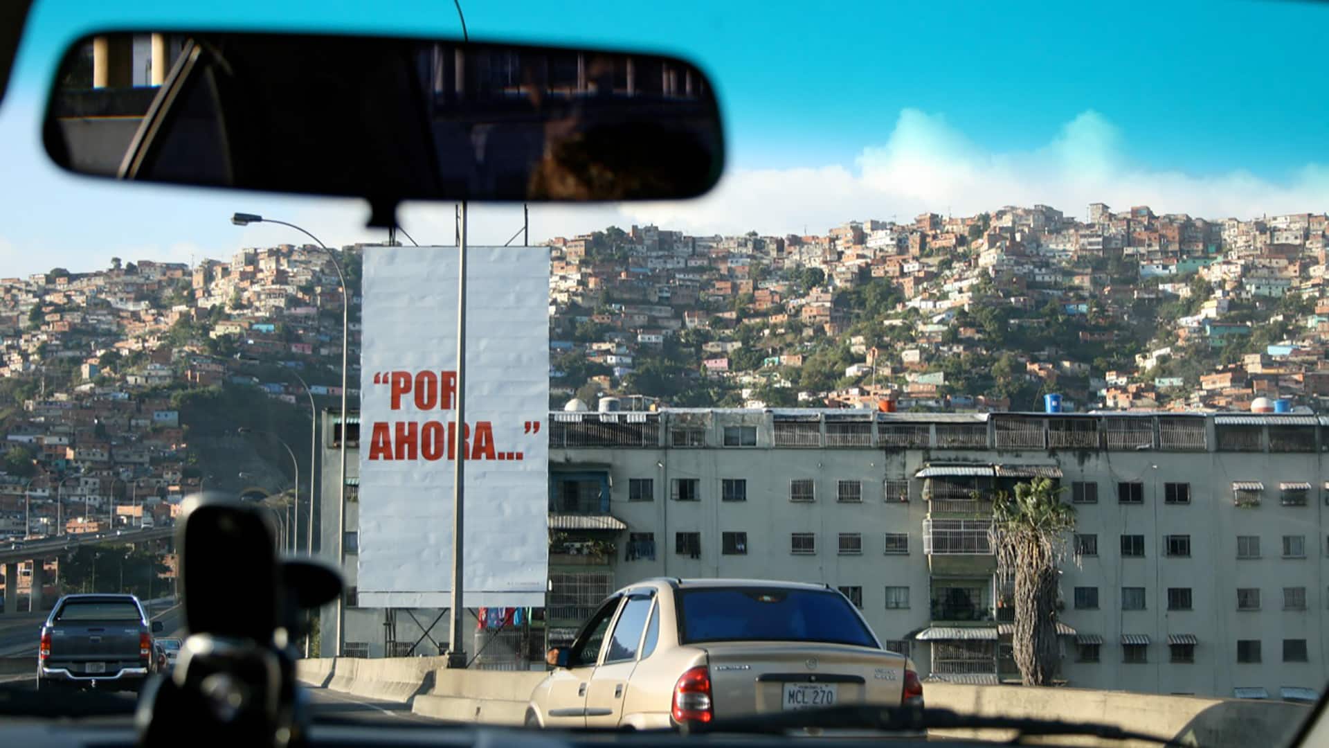 Foto de una pancarta blanca con la frase en rojo “Por ahora…”, tomada desde un coche en la autopista Caracas – La Guaira. Detrás de la pancarta, un edificio bajo de apartamentos y al fondo la imagen de un cerro o barriada popular en el oeste de la ciudad de Caracas (Venezuela). La frase fue pronunciada por Hugo Chávez Frías en su rendición ante los medios de comunicación, tras el intento de golpe de Estado en 1992.