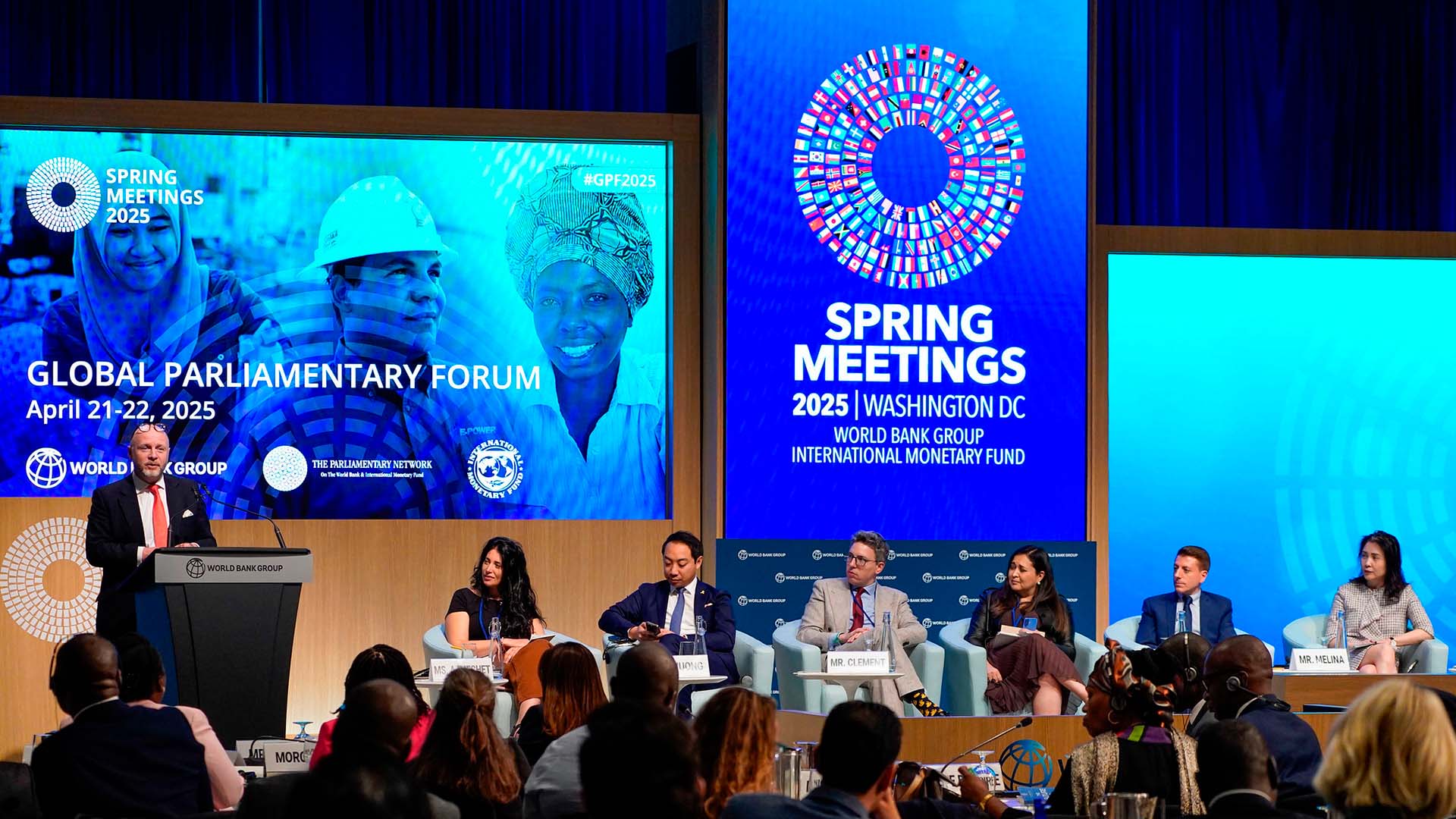 Panel internacional en el Global Parliamentary Forum 2025 durante las Reuniones de Primavera del Banco Mundial y el FMI en Washington D.C. A la izquierda, de pie, un hombre modera el panel desde el podio, mientras seis panelistas debaten frente a una audiencia diversa. Las pantallas LED muestran logotipos del evento y rostros de diferentes personas. Reuniones de primavera