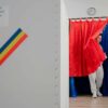 A voter steps out of a booth draped in Romania’s red, yellow, and blue during the first round of the 2025 presidential election, with a ballot box marked 'Alegerea Președintelui României 2025' in view. Romania