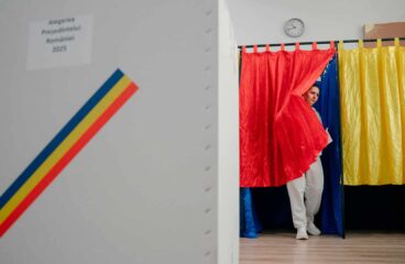 A voter steps out of a booth draped in Romania’s red, yellow, and blue during the first round of the 2025 presidential election, with a ballot box marked 'Alegerea Președintelui României 2025' in view. Romania