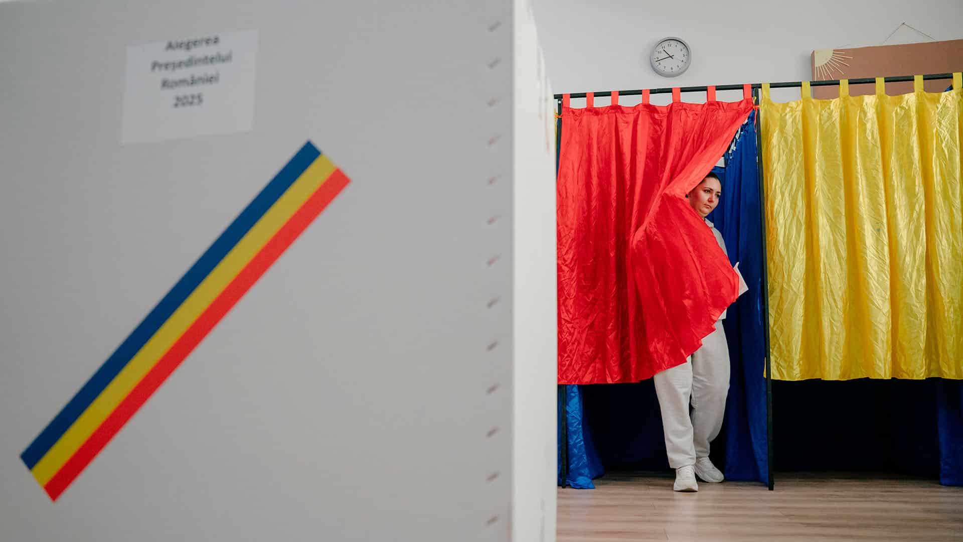 A voter steps out of a booth draped in Romania’s red, yellow, and blue during the first round of the 2025 presidential election, with a ballot box marked 'Alegerea Președintelui României 2025' in view. Romania
