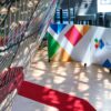 Vista interior del edificio Europa en Bruselas durante la Cumbre UE-CELAC 2023. Destaca una alfombra roja en zigzag sobre el suelo blanco, una pared con el logotipo multicolor del evento, y filas de banderas nacionales ubicadas a la derecha. También se aprecian patrones de luz solar filtrados a través de la fachada de cristal y metal. UE-CELAC