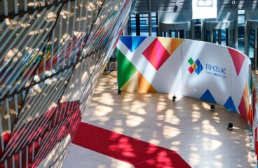 Vista interior del edificio Europa en Bruselas durante la Cumbre UE-CELAC 2023. Destaca una alfombra roja en zigzag sobre el suelo blanco, una pared con el logotipo multicolor del evento, y filas de banderas nacionales ubicadas a la derecha. También se aprecian patrones de luz solar filtrados a través de la fachada de cristal y metal. UE-CELAC