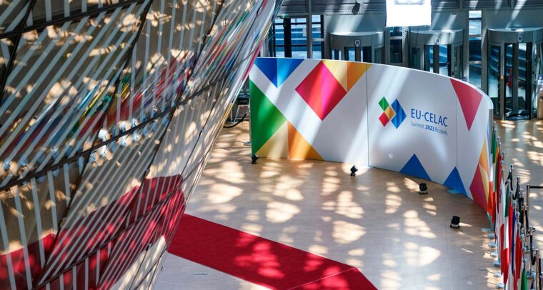 Vista interior del edificio Europa en Bruselas durante la Cumbre UE-CELAC 2023. Destaca una alfombra roja en zigzag sobre el suelo blanco, una pared con el logotipo multicolor del evento, y filas de banderas nacionales ubicadas a la derecha. También se aprecian patrones de luz solar filtrados a través de la fachada de cristal y metal. UE-CELAC