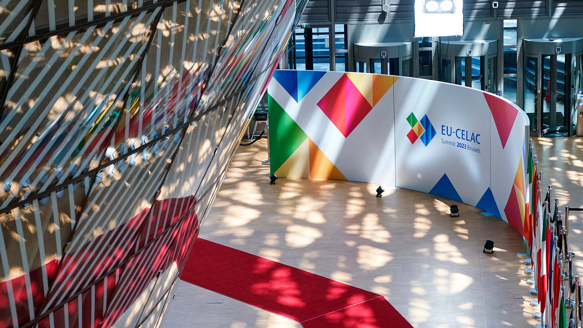 Vista interior del edificio Europa en Bruselas durante la Cumbre UE-CELAC 2023. Destaca una alfombra roja en zigzag sobre el suelo blanco, una pared con el logotipo multicolor del evento, y filas de banderas nacionales ubicadas a la derecha. También se aprecian patrones de luz solar filtrados a través de la fachada de cristal y metal. UE-CELAC