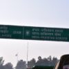 Primer plano de una señal de tráfico verde que indica la distancia a Lahore (23 km) y a la frontera entre India y Pakistán (1 km), en panyabí, hindi e inglés. A la izquierda y al fondo se avistan árboles, y en el fondo se aprecia una bandera de Pakistán. Cachemira