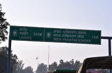 Primer plano de una señal de tráfico verde que indica la distancia a Lahore (23 km) y a la frontera entre India y Pakistán (1 km), en panyabí, hindi e inglés. A la izquierda y al fondo se avistan árboles, y en el fondo se aprecia una bandera de Pakistán. Cachemira
