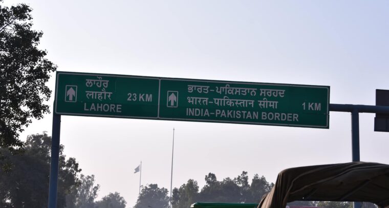 Primer plano de una señal de tráfico verde que indica la distancia a Lahore (23 km) y a la frontera entre India y Pakistán (1 km), en panyabí, hindi e inglés. A la izquierda y al fondo se avistan árboles, y en el fondo se aprecia una bandera de Pakistán. Cachemira