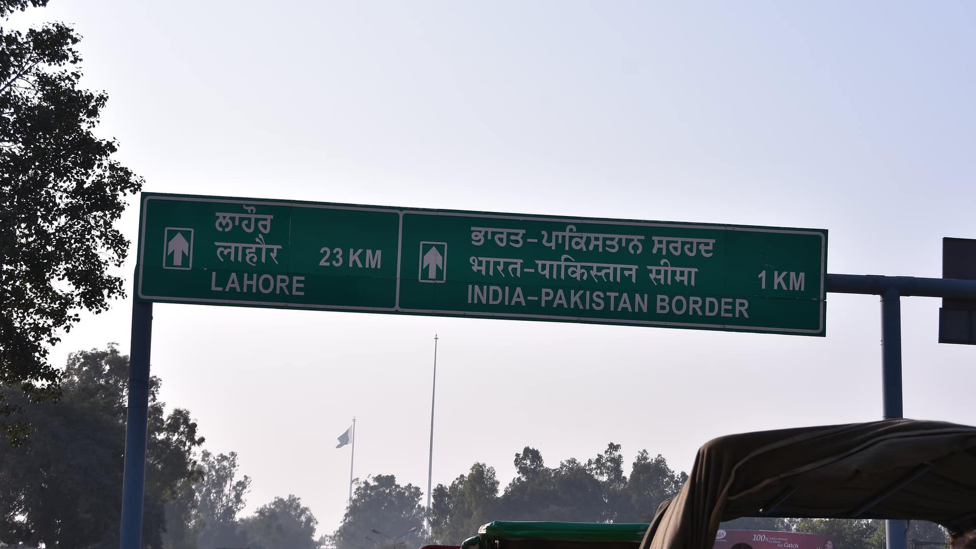 Primer plano de una señal de tráfico verde que indica la distancia a Lahore (23 km) y a la frontera entre India y Pakistán (1 km), en panyabí, hindi e inglés. A la izquierda y al fondo se avistan árboles, y en el fondo se aprecia una bandera de Pakistán. Cachemira