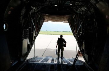 Vista desde el interior de una aeronave militar de transporte, con un soldado descendiendo por la rampa trasera hacia la pista de aterrizaje en un día soleado. UE