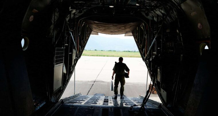 Vista desde el interior de una aeronave militar de transporte, con un soldado descendiendo por la rampa trasera hacia la pista de aterrizaje en un día soleado. UE