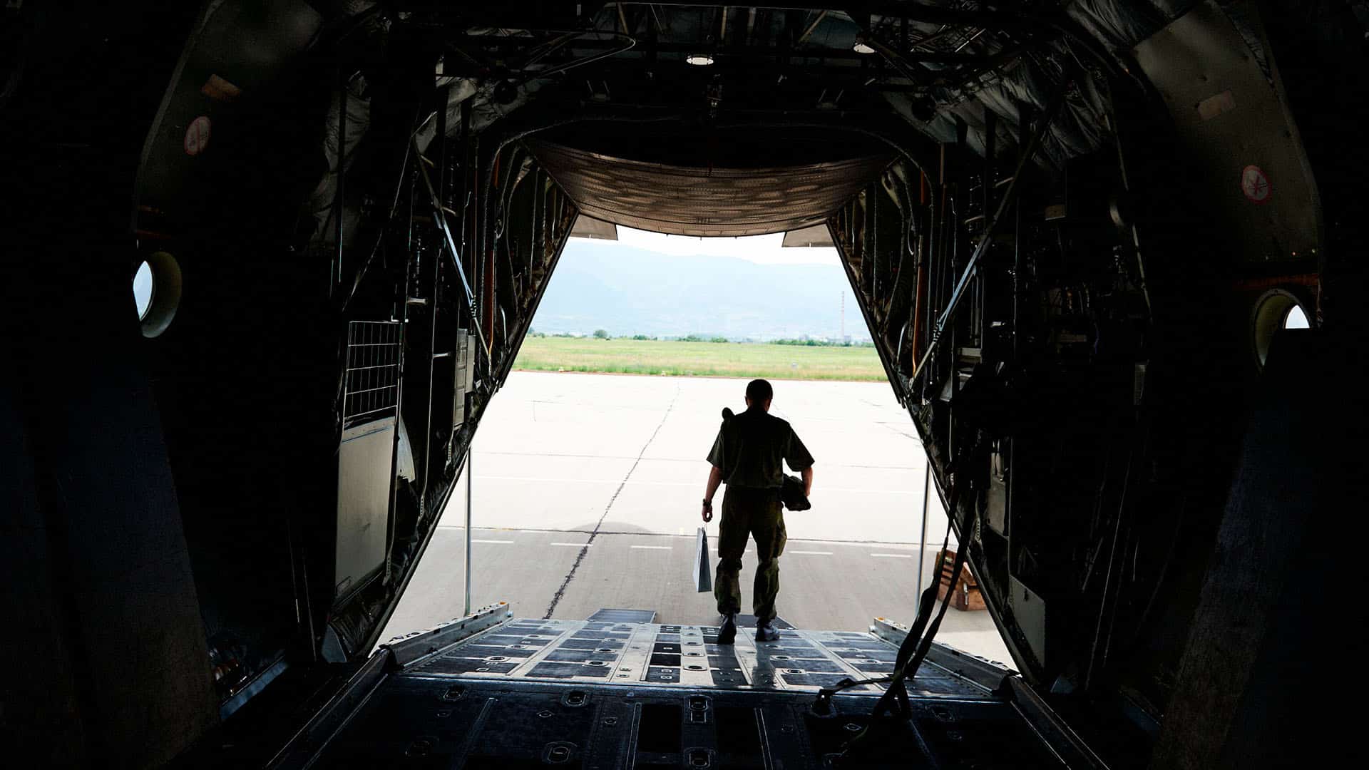 Vista desde el interior de una aeronave militar de transporte, con un soldado descendiendo por la rampa trasera hacia la pista de aterrizaje en un día soleado. UE