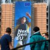Three people stand in front of the Berlaymont building in Brussels, where a large vertical banner for the EU’s NextGenerationEU programme shows a young woman stepping forward, with “Next Gen EU” and the EU flag above her. NGEU