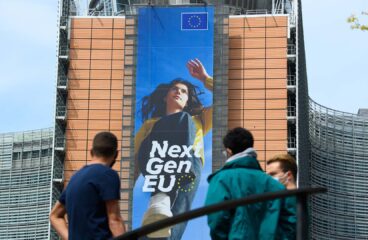Three people stand in front of the Berlaymont building in Brussels, where a large vertical banner for the EU’s NextGenerationEU programme shows a young woman stepping forward, with “Next Gen EU” and the EU flag above her. NGEU