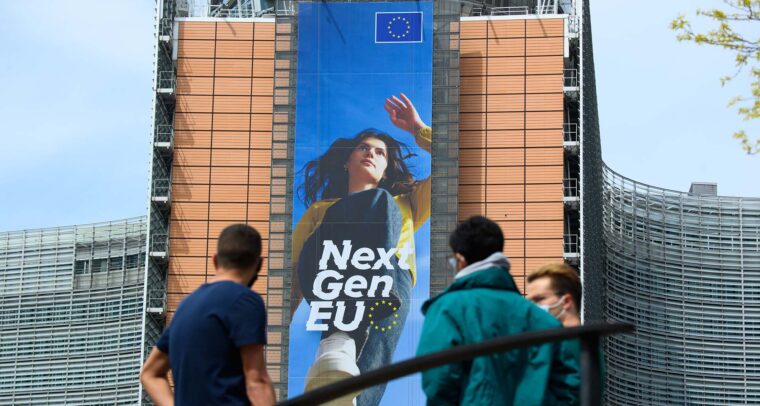 Three people stand in front of the Berlaymont building in Brussels, where a large vertical banner for the EU’s NextGenerationEU programme shows a young woman stepping forward, with “Next Gen EU” and the EU flag above her. NGEU