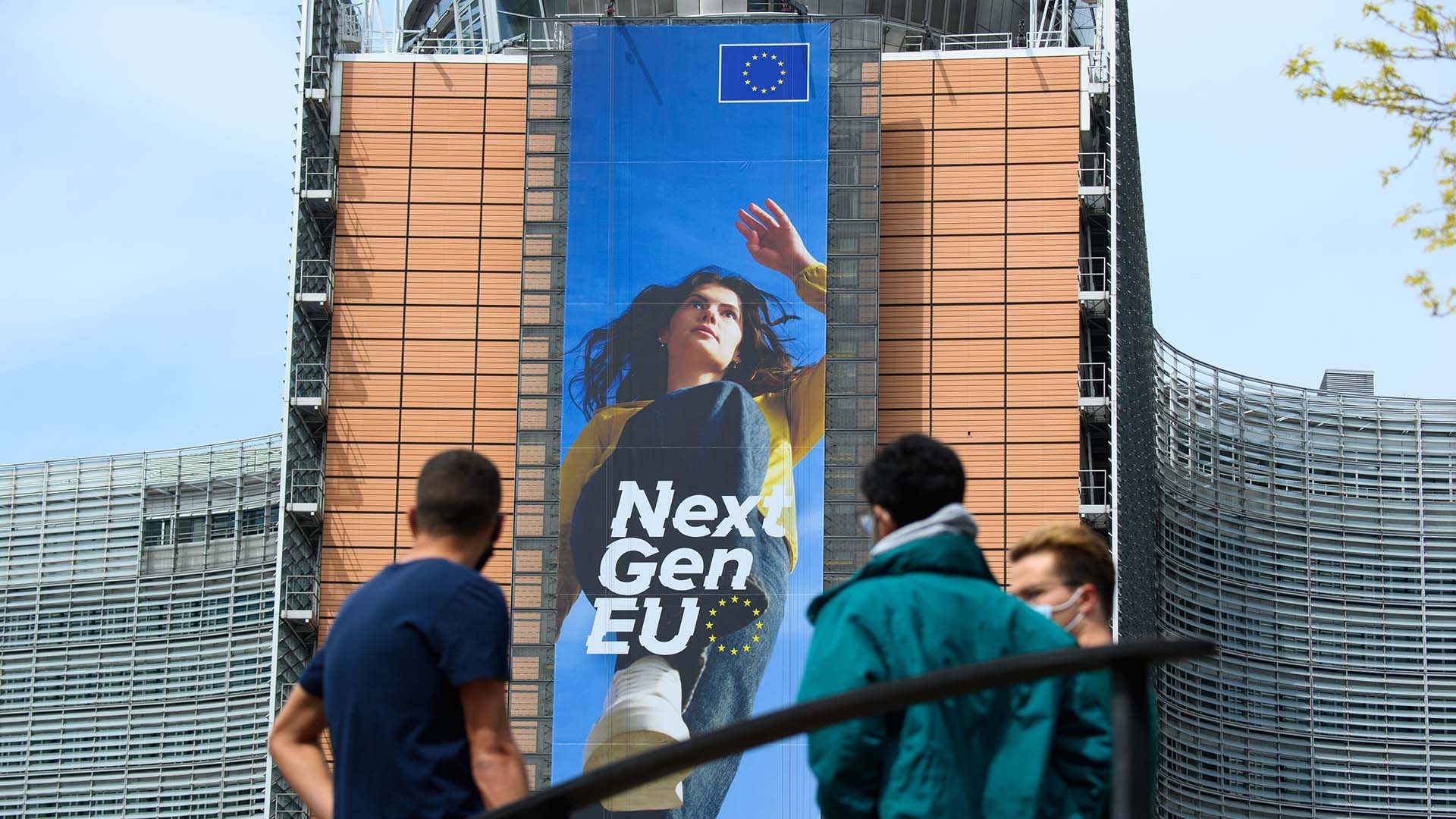 Three people stand in front of the Berlaymont building in Brussels, where a large vertical banner for the EU’s NextGenerationEU programme shows a young woman stepping forward, with “Next Gen EU” and the EU flag above her. NGEU