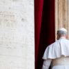 Pope Leo IV, seen from behind and dressed in the papal cassock, walked away after delivering his first Regina Caeli prayer from a balcony overlooking St. Peter’s Square. On the left, part of the building clad in light travertine can be seen, while a column of the same stone, framed by red curtains, stands behind him. Pope