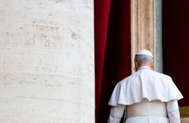 Pope Leo IV, seen from behind and dressed in the papal cassock, walked away after delivering his first Regina Caeli prayer from a balcony overlooking St. Peter’s Square. On the left, part of the building clad in light travertine can be seen, while a column of the same stone, framed by red curtains, stands behind him. Pope