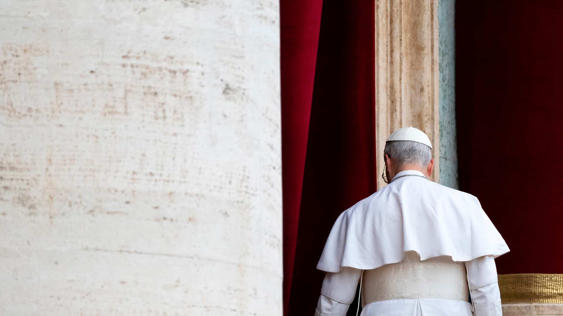 Pope Leo IV, seen from behind and dressed in the papal cassock, walked away after delivering his first Regina Caeli prayer from a balcony overlooking St. Peter’s Square. On the left, part of the building clad in light travertine can be seen, while a column of the same stone, framed by red curtains, stands behind him. Pope