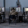 Sala de instrucción militar, con tres soldados sentados (con uniformes de camuflaje y auriculares) frente a simuladores de tanques Leopard. La sala, del centro de entrenamiento en Świętoszów (Polonia), tiene paredes metálicas, suelo de hormigón y estaciones de simulación alineadas para el entrenamiento técnico. Ucrania