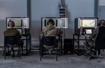 Sala de instrucción militar, con tres soldados sentados (con uniformes de camuflaje y auriculares) frente a simuladores de tanques Leopard. La sala, del centro de entrenamiento en Świętoszów (Polonia), tiene paredes metálicas, suelo de hormigón y estaciones de simulación alineadas para el entrenamiento técnico. Ucrania