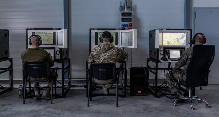 Sala de instrucción militar, con tres soldados sentados (con uniformes de camuflaje y auriculares) frente a simuladores de tanques Leopard. La sala, del centro de entrenamiento en Świętoszów (Polonia), tiene paredes metálicas, suelo de hormigón y estaciones de simulación alineadas para el entrenamiento técnico. Ucrania