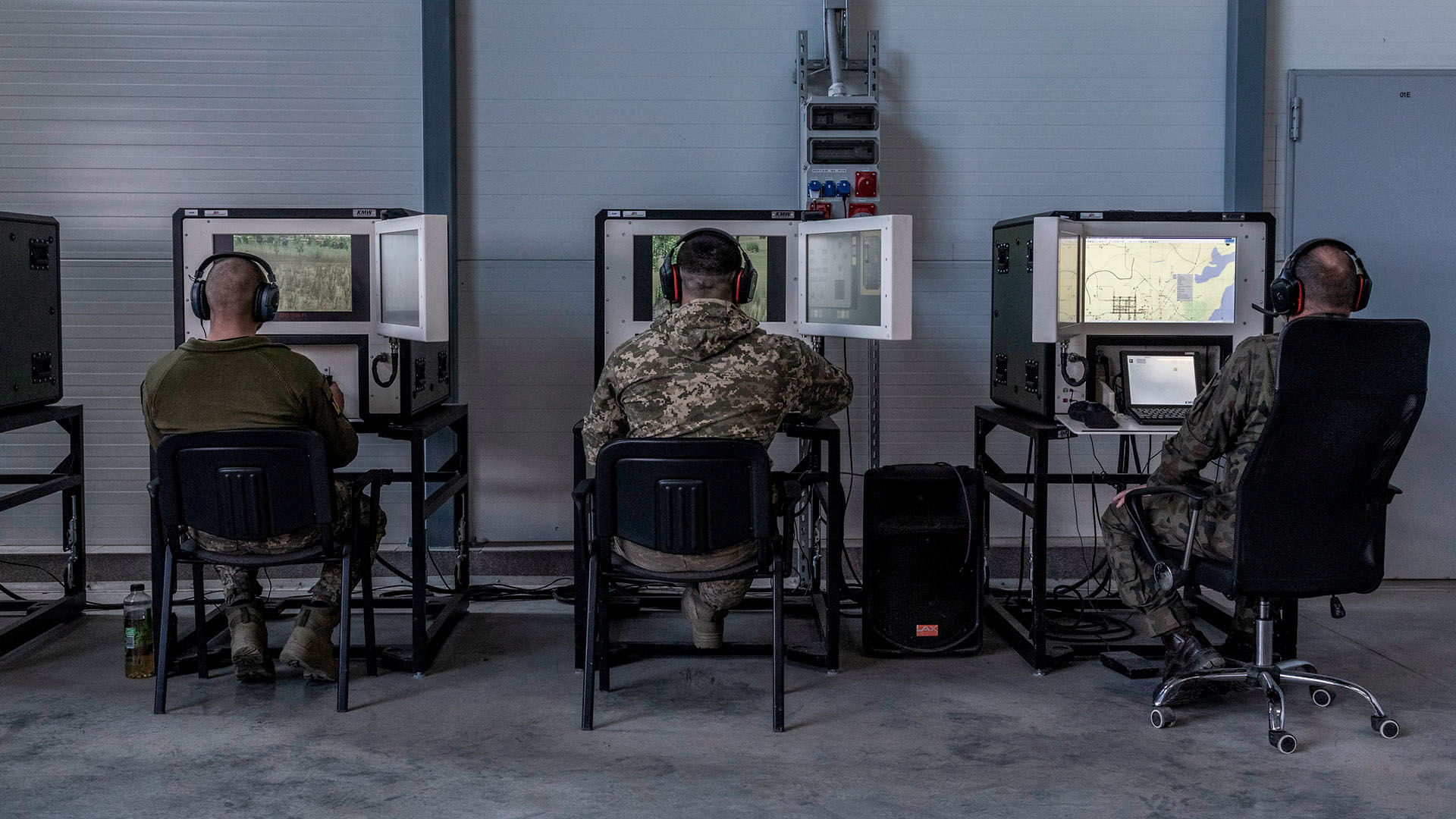 Sala de instrucción militar, con tres soldados sentados (con uniformes de camuflaje y auriculares) frente a simuladores de tanques Leopard. La sala, del centro de entrenamiento en Świętoszów (Polonia), tiene paredes metálicas, suelo de hormigón y estaciones de simulación alineadas para el entrenamiento técnico. Ucrania