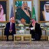 Donald Trump y el príncipe heredero saudí Mohammed bin Salman conversan sentados en una sala lujosamente decorada en Riad, con retratos de líderes saudíes al fondo, banderas de EEUU y Arabia Saudí tras ellos, y mesas doradas con arreglos florales. Trump