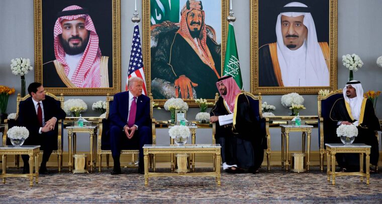 Donald Trump y el príncipe heredero saudí Mohammed bin Salman conversan sentados en una sala lujosamente decorada en Riad, con retratos de líderes saudíes al fondo, banderas de EEUU y Arabia Saudí tras ellos, y mesas doradas con arreglos florales. Trump